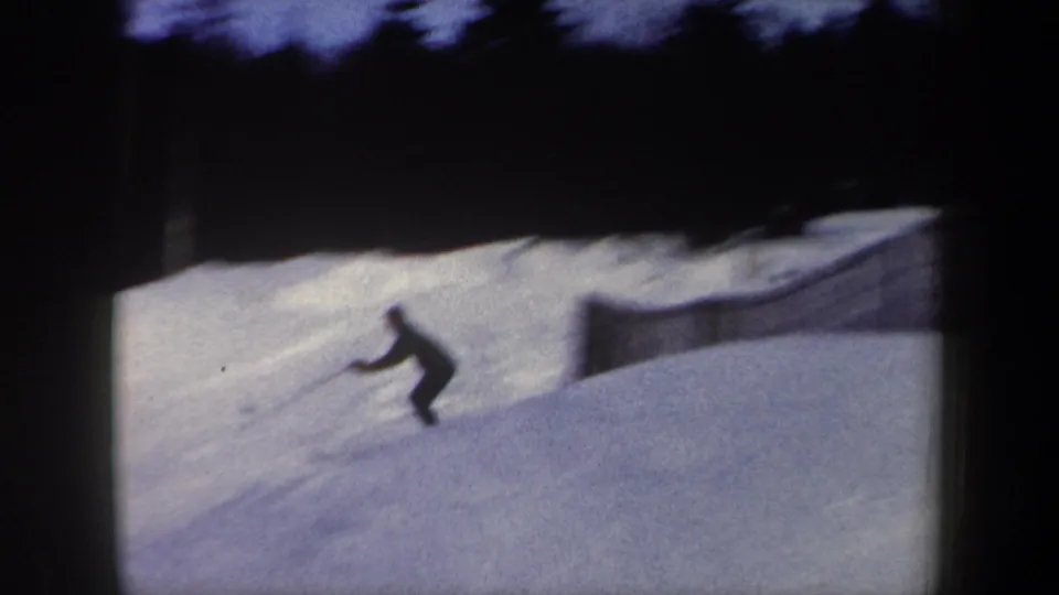 A skier in dark clothing turns across a snowy downhill run near a fence, with dark trees and a bright snowbank behind them in the New York home-movie footage.