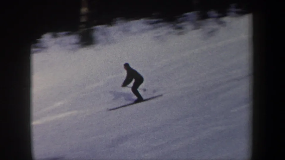 The home-movie frame shows a skier in a low stance crossing the snow, with long skis visible against a softly exposed winter slope.
