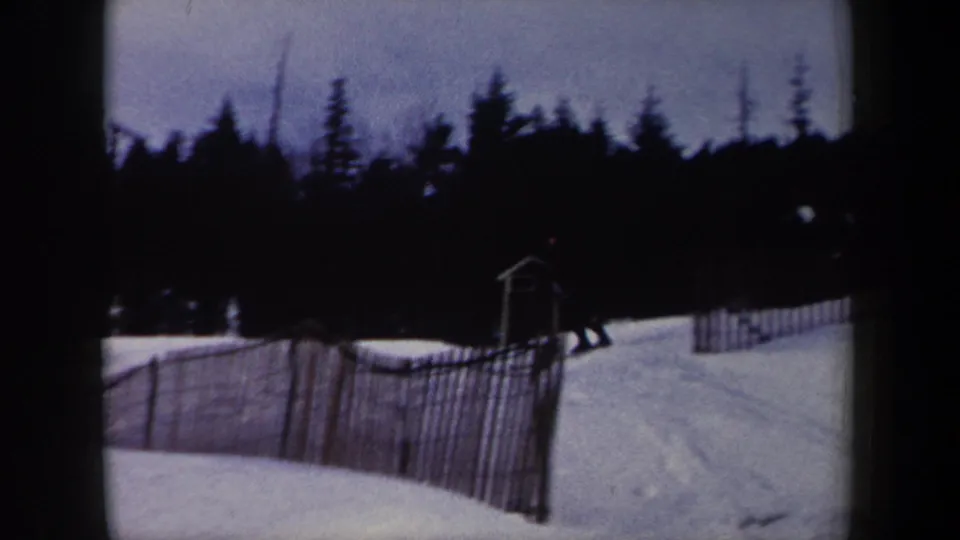 A snowy New York ski slope is seen with slatted fencing, a dark evergreen tree line, and a small skier shape near the right side of the frame.