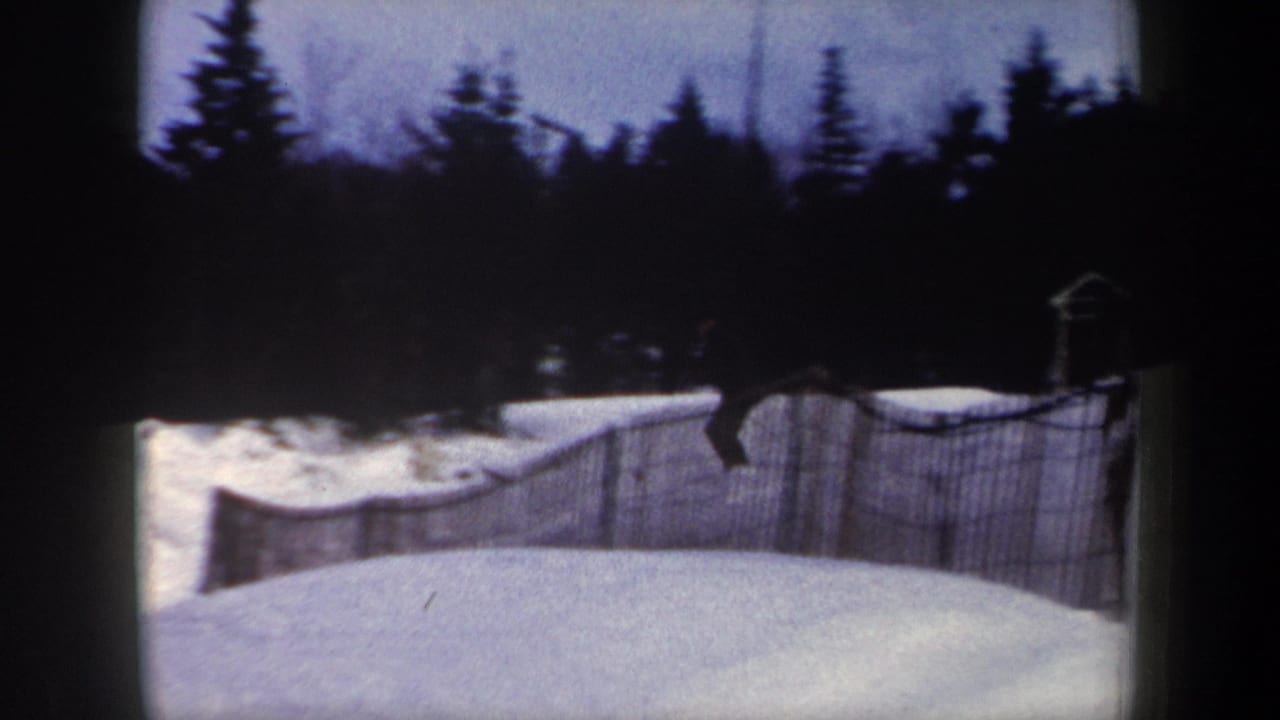 1962 New York home-movie frame showing a snowy hill, wood snow fence, and dark evergreens before the skier enters the run.