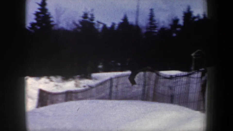 1962 New York home-movie frame showing a snowy hill, wood snow fence, and dark evergreens before the skier enters the run.