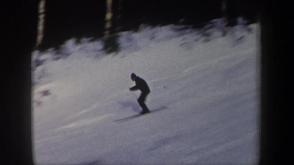 The skier continues downhill across the snow, leaning slightly with poles visible in the soft, grainy 8mm frame.