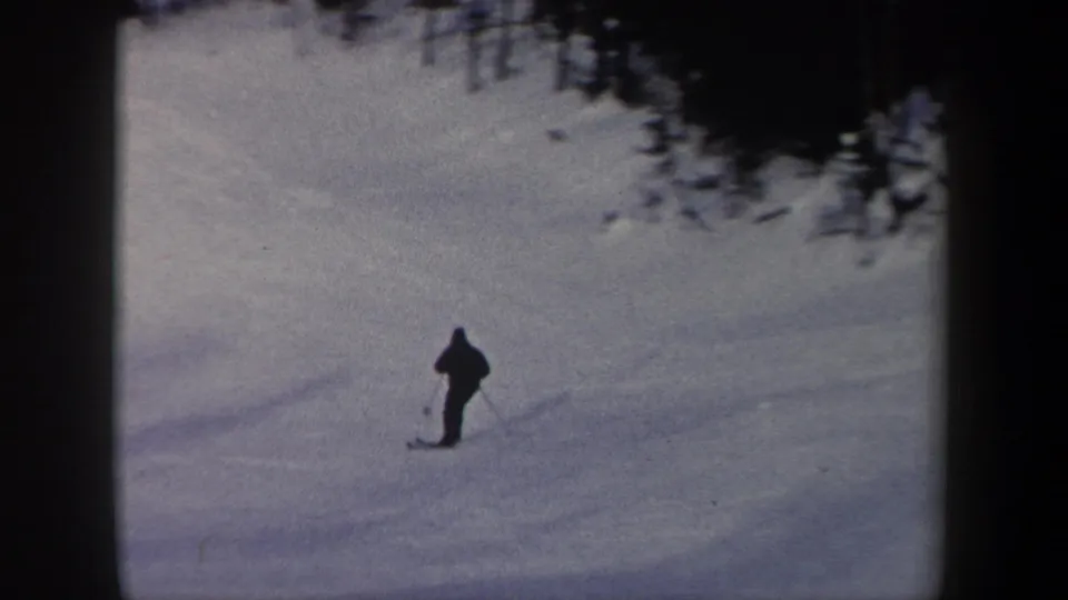 A small figure on skis moves across the lower snowy slope, with dark evergreens above and muted 1960s home-movie color.