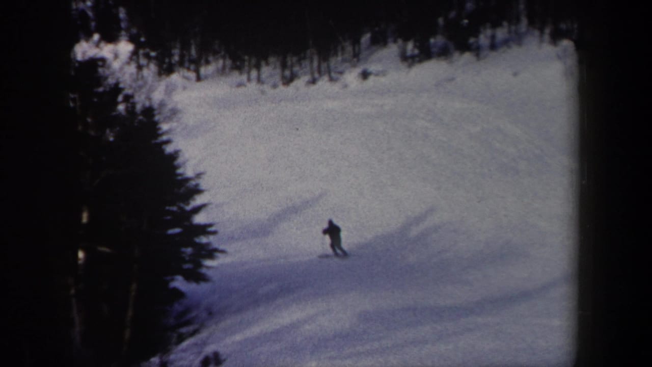 The skier appears small against a broad snowy hillside, with dark trees framing the upper edge of the 8mm frame.