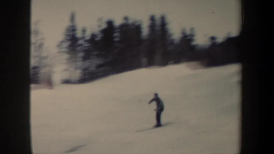 A distant skier in dark clothing moves over a bright snow-covered slope, with evergreen trees along the background.