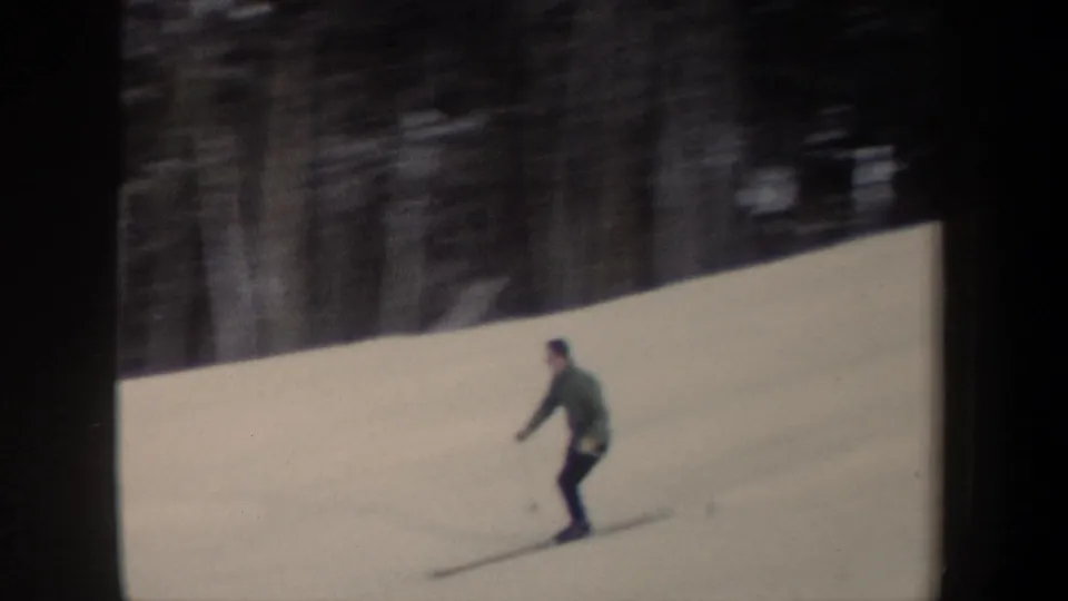 1962 New York home-movie frame with a skier crossing a snow-covered slope, backed by bare trees and evergreens.