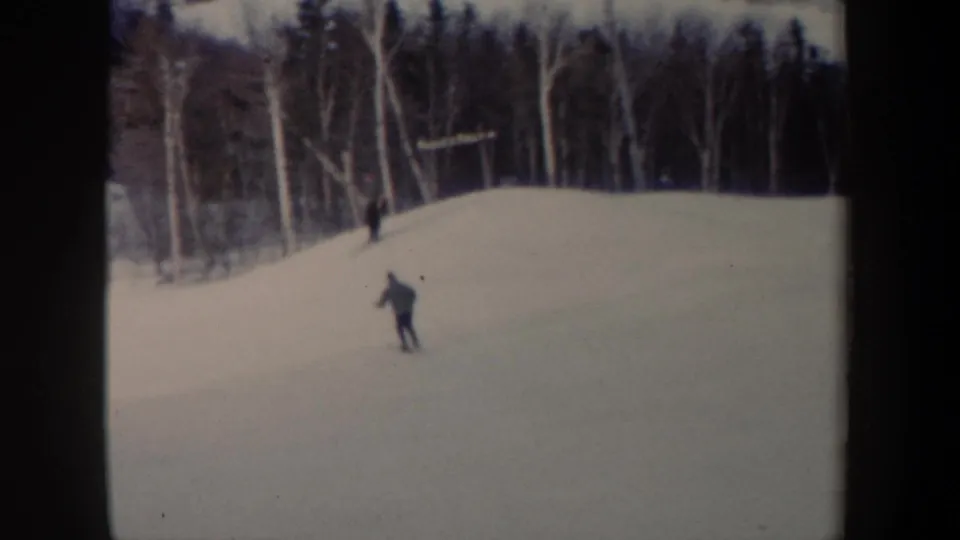 1962 New York home-movie frame showing a skier lower on the snowy hill, with a distant figure near the trees above.