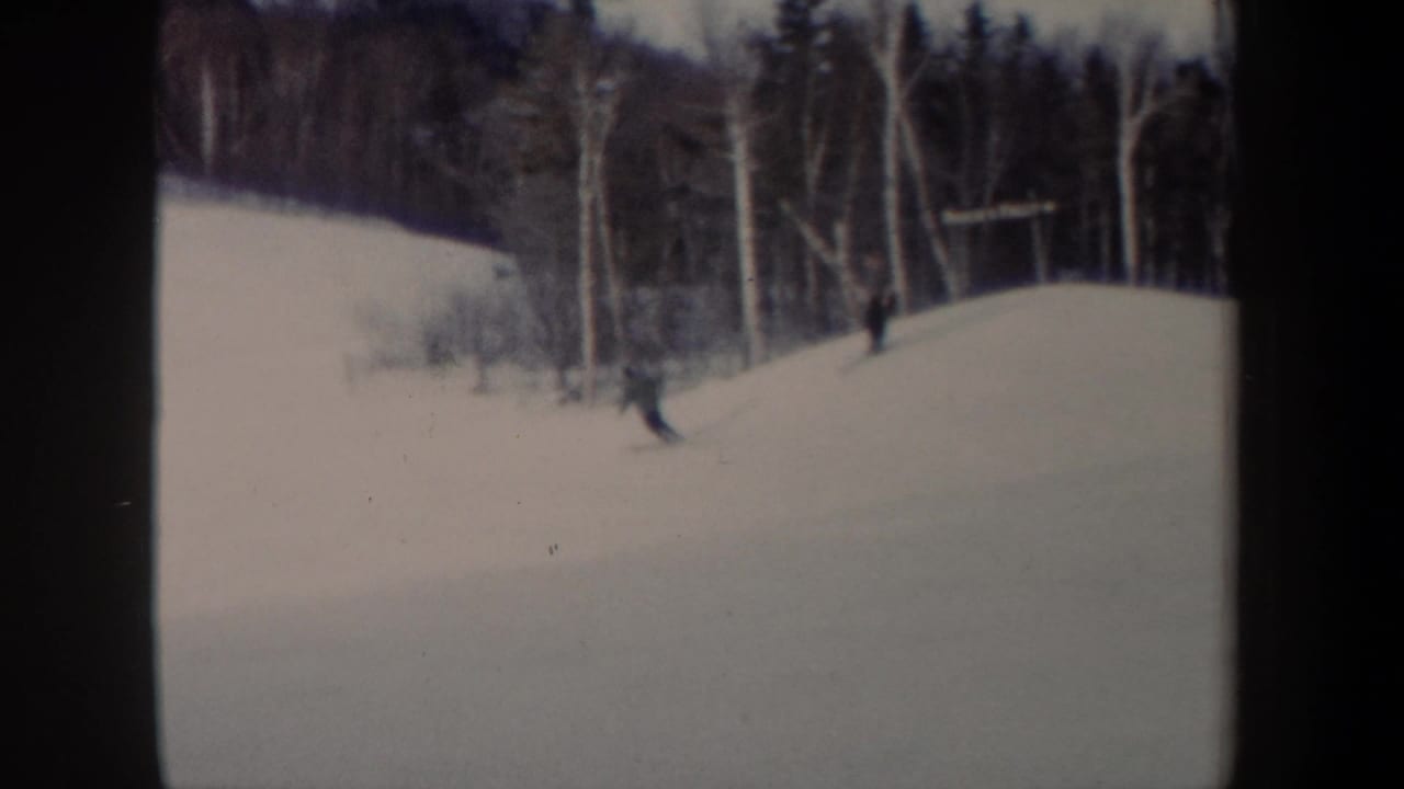 1962 New York home-movie frame showing a distant skier moving downhill on a snowy slope with bare trees behind.