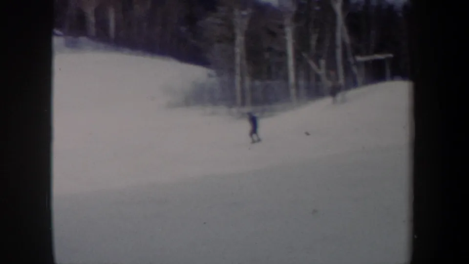 A small figure skis downhill beneath tall winter trees in soft, grainy 8mm footage from New York, 1962.