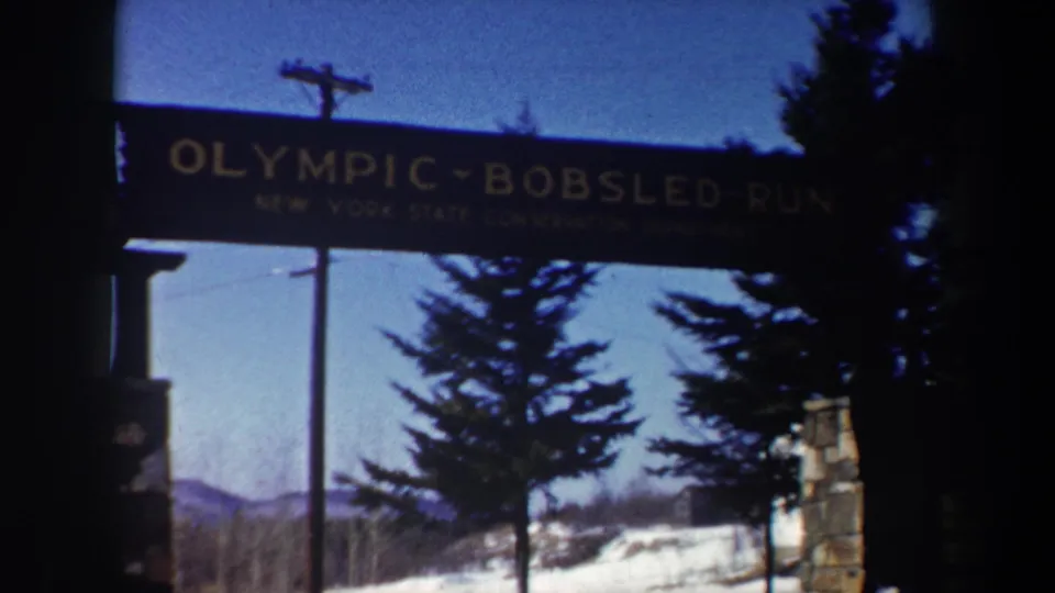 1962 New York 8mm view of the Olympic Bobsled Run entrance, with the sign partly shadowed above evergreens, snow, and a distant ridge.