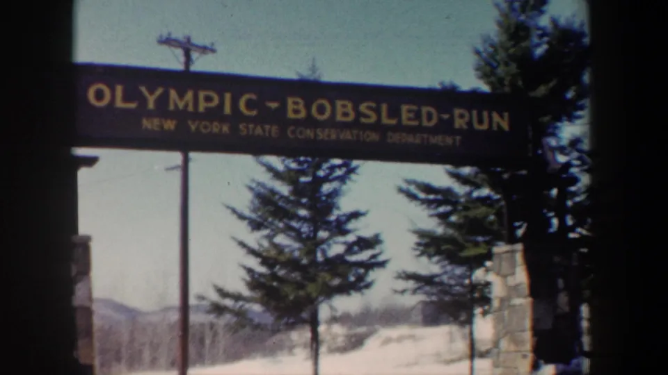 1962 New York home-movie frame showing the Olympic Bobsled Run sign, snowy foreground, evergreens, and stone entrance structure.