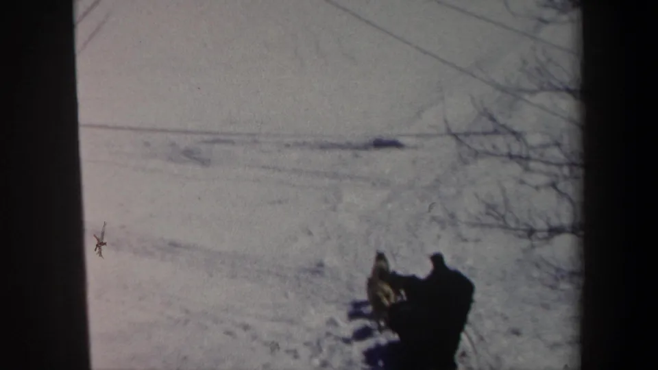 1962 New York 8mm frame showing the sled rider and dog team farther across open snow, with faint tracks and branches visible.