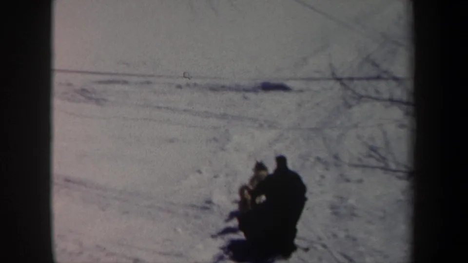 1962 New York 8mm frame showing a distant musher and dog team crossing a snowy field under bare branches.