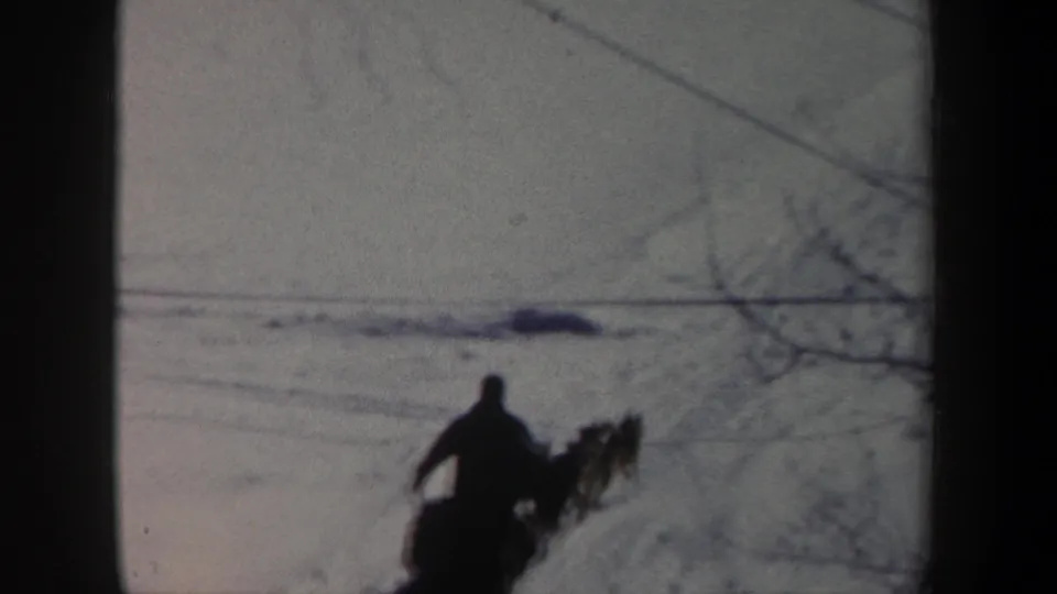 Distant 1962 New York home-movie frame of a dog sled team moving through snow with utility lines overhead.