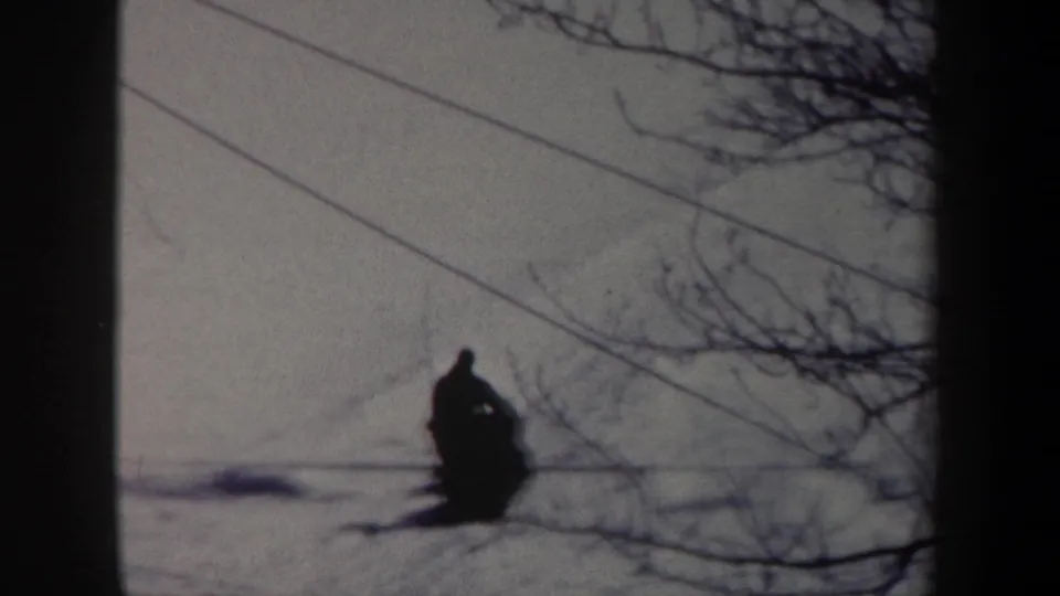 1962 New York 8mm frame showing a distant sled rider moving across a snowy field under bare branches and overhead lines.