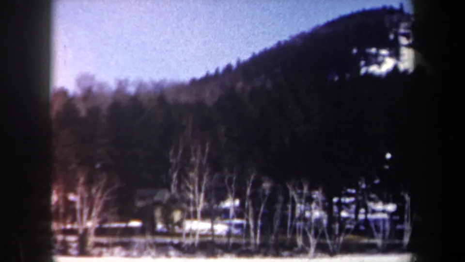 1962 New York 8mm frame of a snowy rural hillside with bare trees, scattered houses, fences, and a dark wooded ridge.