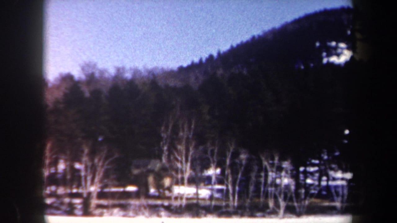 1962 New York 8mm frame showing a winter treeline, dark wooded hillside, pale blue sky, and snow along the ground.