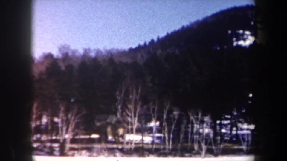 1962 New York 8mm frame showing a winter treeline, dark wooded hillside, pale blue sky, and snow along the ground.