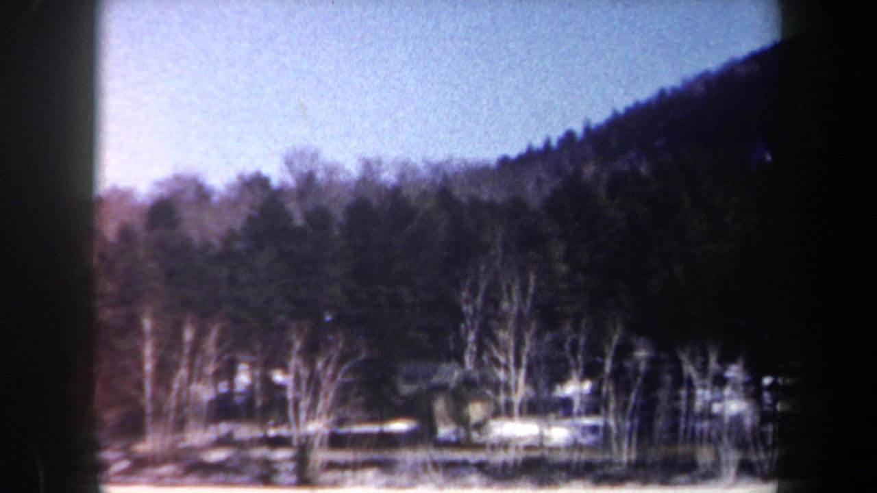 1962 New York 8mm frame showing a snowfield, bare trees, evergreens, hillside, and a distant cabin-like structure.