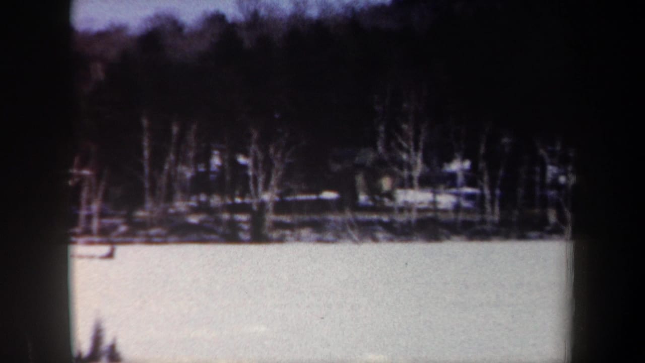 Snowy open ground and treeline with a distant cabin-like structure partly obscured by trees.