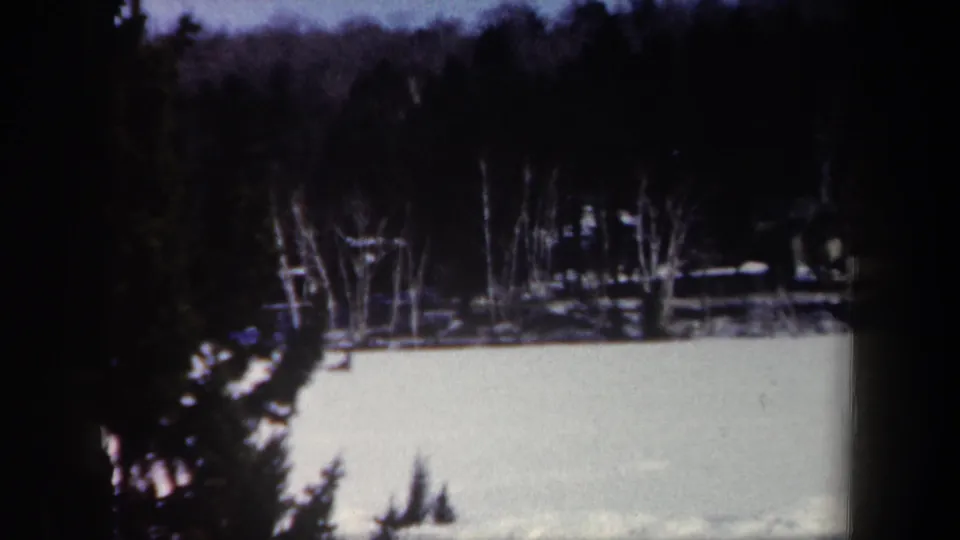 1962 New York 8mm winter frame with a snowy field, wooded hillside, evergreens, and a cabin-like structure through trees.