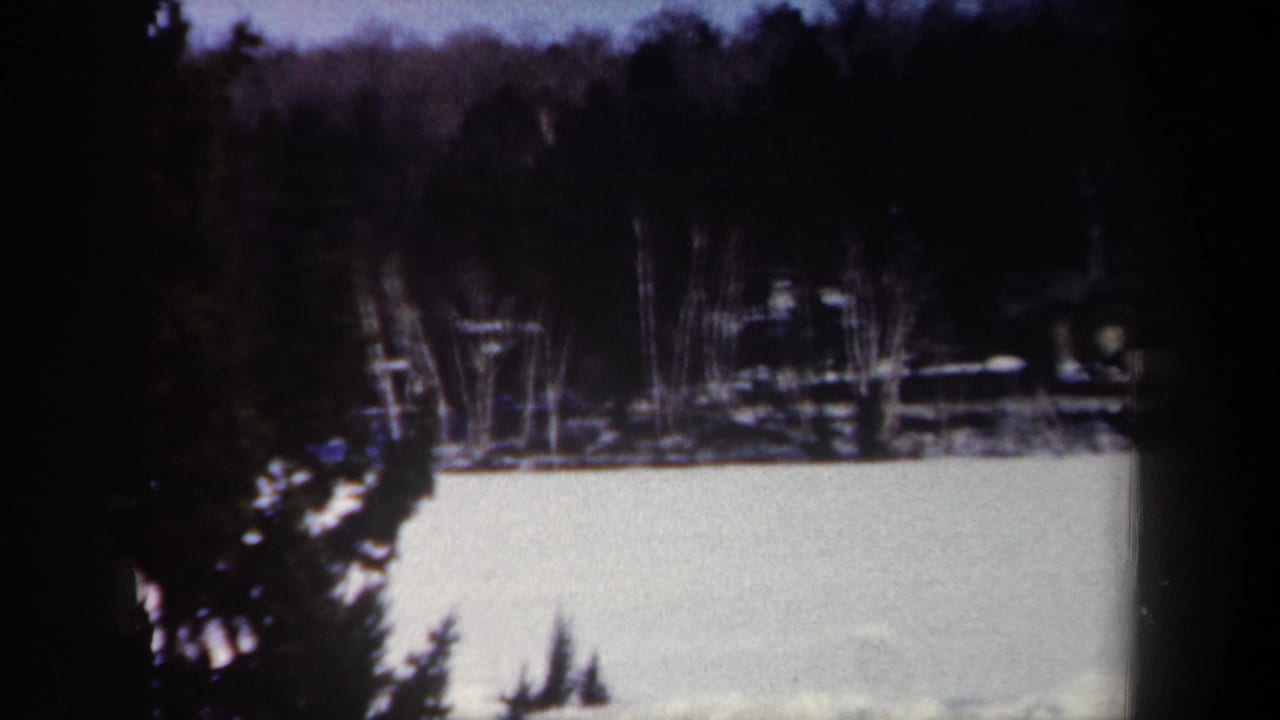 1962 New York 8mm frame showing a pale frozen lake, dark wooded shoreline, and evergreen branches in the foreground.