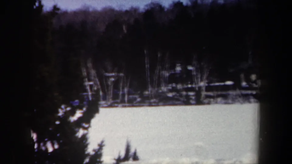 1962 New York 8mm frame showing a pale frozen lake, dark wooded shoreline, and evergreen branches in the foreground.