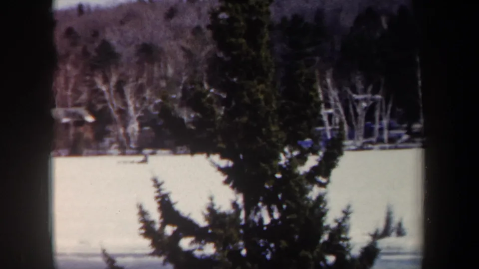 1962 New York 8mm frame showing a snowy frozen lake, evergreen foliage, and a wooded shoreline in winter.