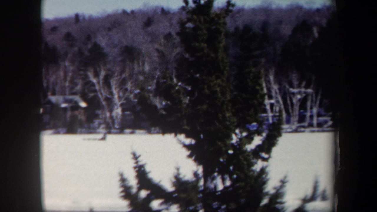 1962 New York 8mm frame showing a snow-covered hill, bare trees, evergreens, and a distant ski jump structure partly obscured by framing.