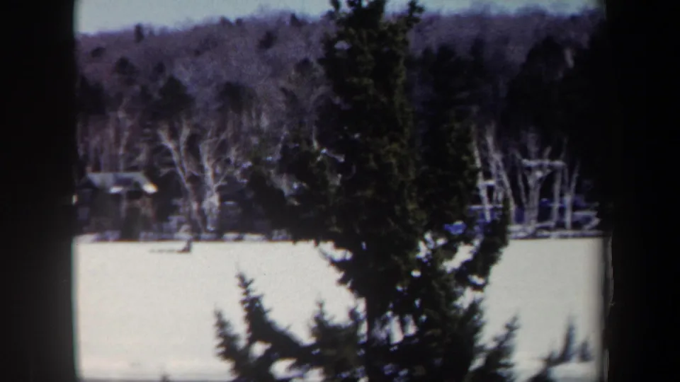 1962 New York 8mm frame showing a snow-covered hill, bare trees, evergreens, and a distant ski jump structure partly obscured by framing.