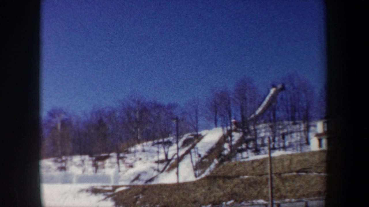 1962 New York 8mm frame with a snow-covered ski jump ramp, bare trees, hillside, and deep blue winter sky.