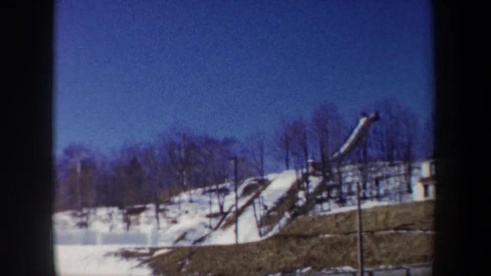 1962 New York 8mm frame with a snow-covered ski jump ramp, bare trees, hillside, and deep blue winter sky.