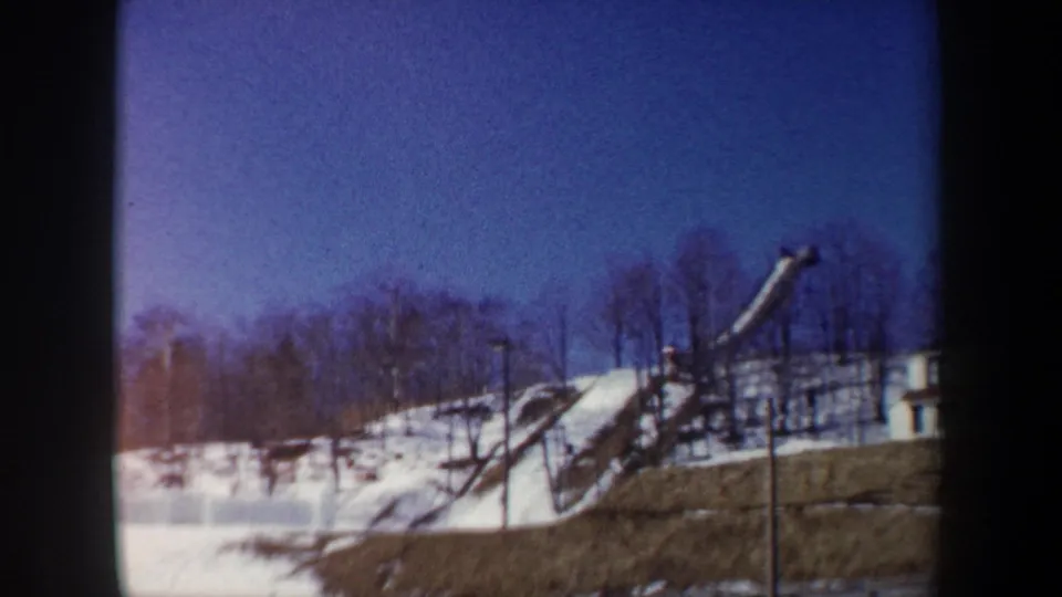1962 New York 8mm frame showing a distant ski jump, snowy hillside, bare winter trees, and open blue sky.