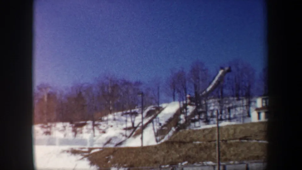 1962 New York 8mm frame of a ski jump ramp on a snowy hill, with bare trees, blue sky, and soft color-shifted film texture.
