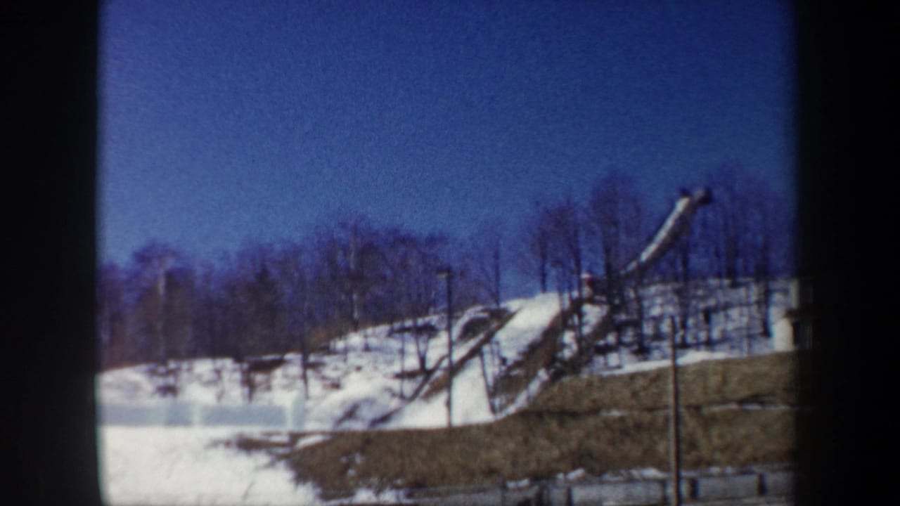 1962 New York home-movie frame of a wooden ski jump ramp set into a snowy slope with leafless trees behind it.