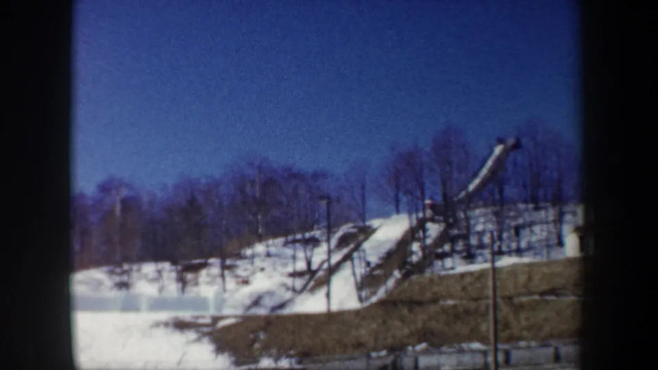 1962 New York home-movie frame of a wooden ski jump ramp set into a snowy slope with leafless trees behind it.