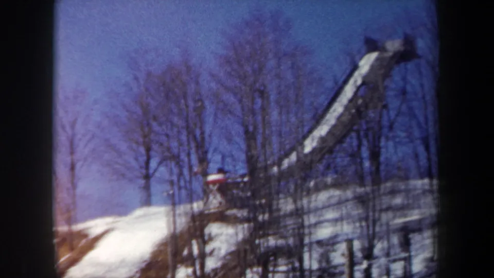1962 New York home-movie frame with a distant winter-clad figure near the top of a snowy ski jump ramp.
