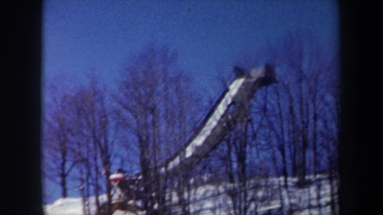 Snow-covered ski jump ramp rises above a wooded winter hillside under a blue sky.