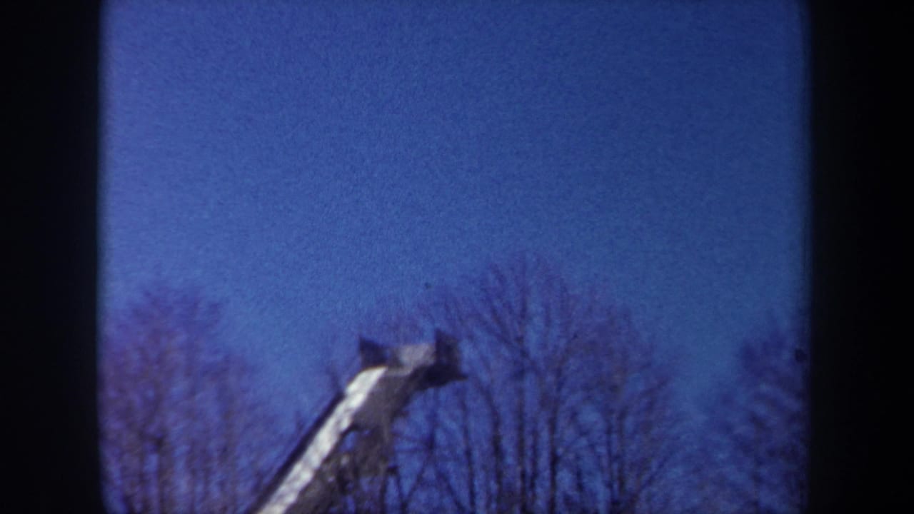 1962 New York home-movie frame with the upper part of a snow-covered ski jump ramp against blue sky and trees.