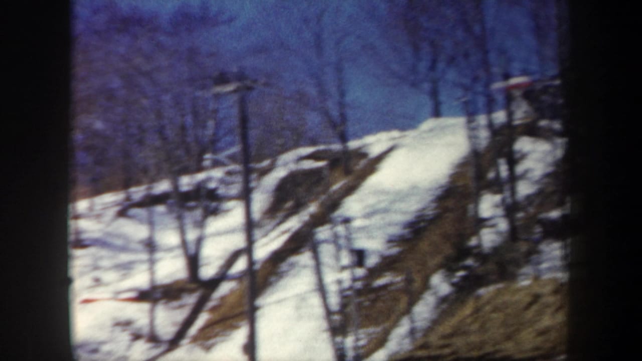 1962 New York 8mm frame showing a snowy hillside, bare trees, lift structures, and a low snow-block wall in the foreground.