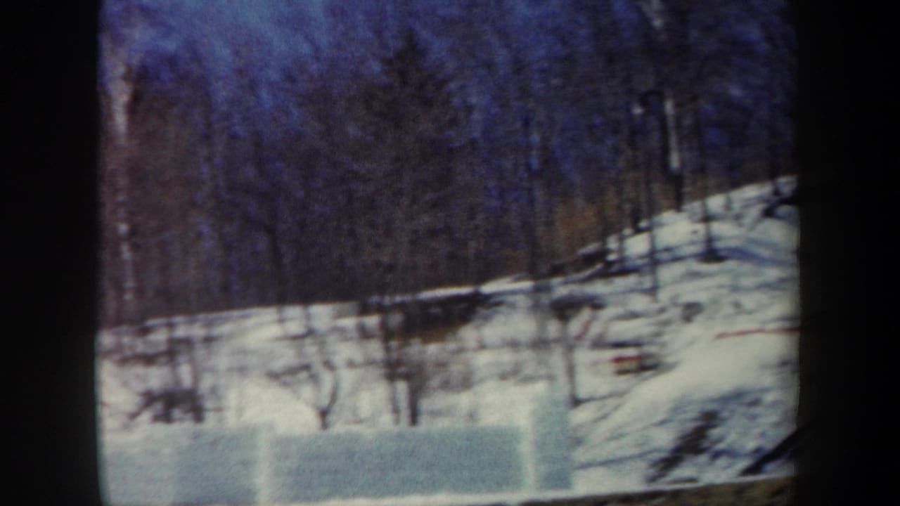 1962 New York winter frame with a low snow-block wall, wooded slope, and possible ski-lift supports under a cold blue sky.