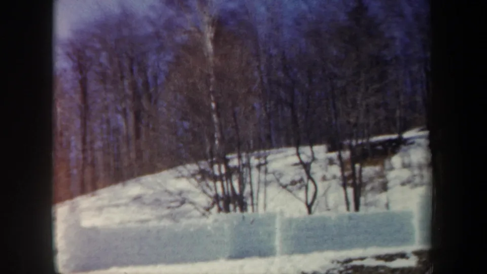 1962 New York 8mm frame showing bare winter trees on a snow-covered hillside behind a low wall of packed snow blocks.