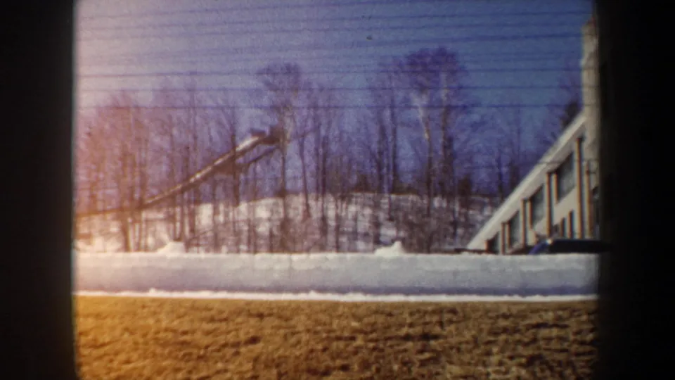 1962 New York winter frame with a snow-block wall across the scene, bare trees on the slope, brown ground, and a building edge at right.