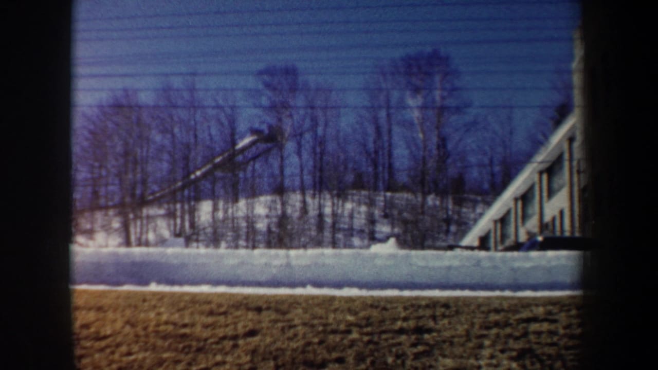 1962 New York 8mm frame showing snow, leafless trees, winter grass, and part of a long building through dark foreground borders.