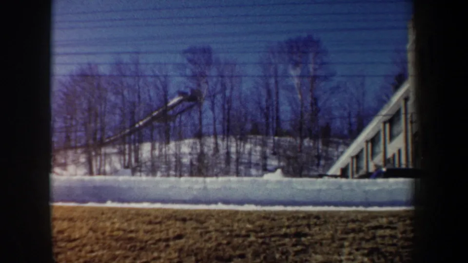 1962 New York 8mm frame showing snow, leafless trees, winter grass, and part of a long building through dark foreground borders.