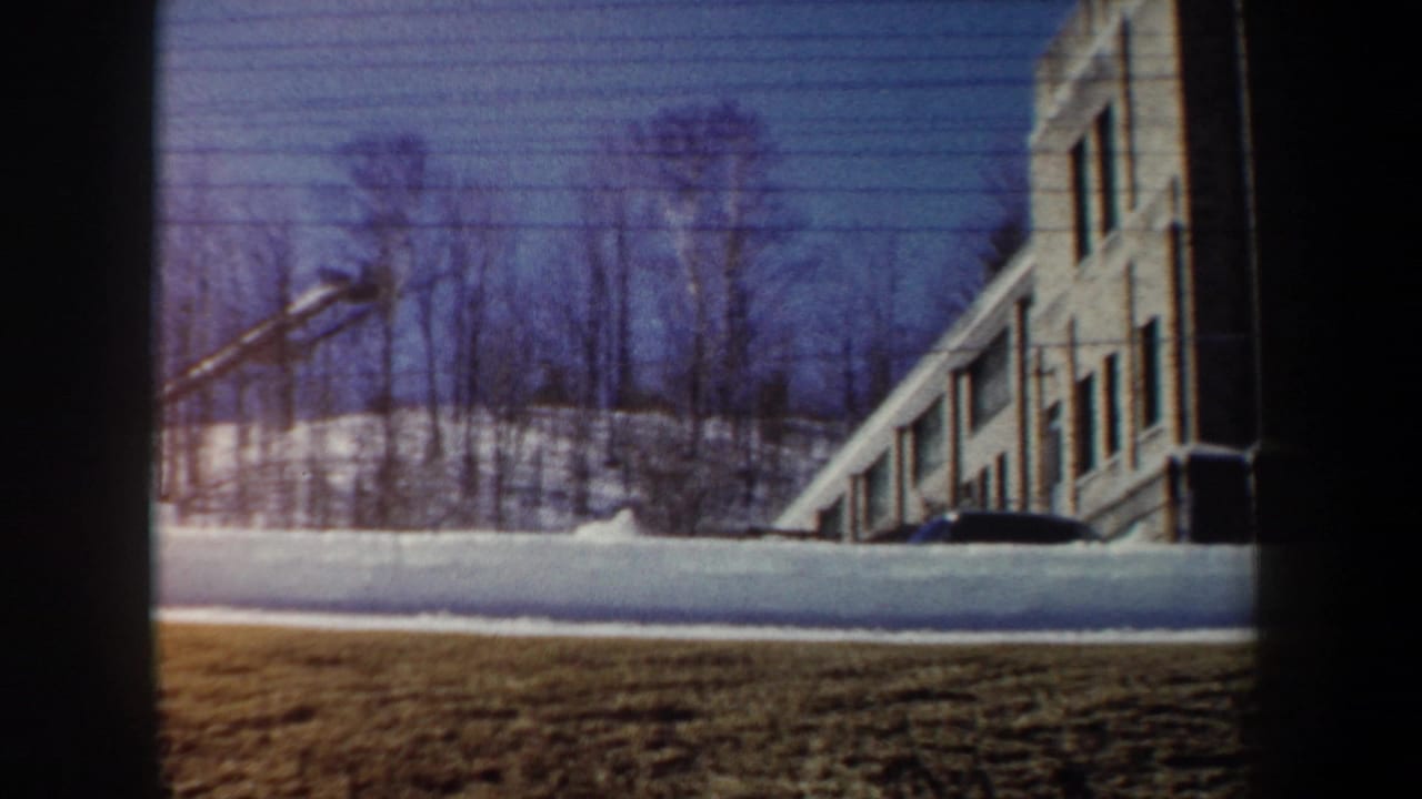 1962 New York winter frame with a snowbank, leafless trees, a windowed building, and a dark vehicle near the wall.