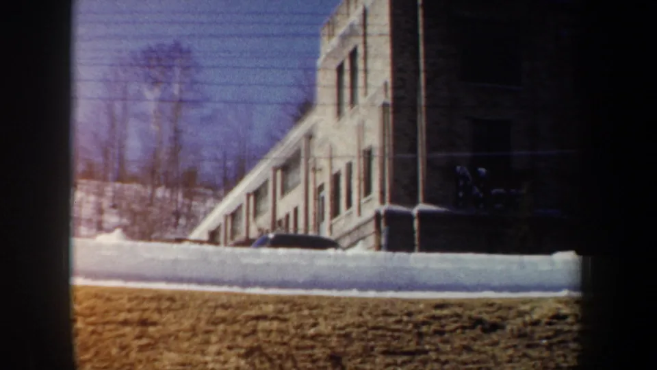 1962 New York 8mm view of a snowbank beside a long building, with leafless trees and dark framing from the camera position.