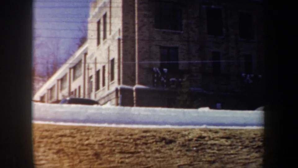 1962 New York winter frame showing a long multi-window building, snow piled along the ground, a dark vehicle, and bare trees.