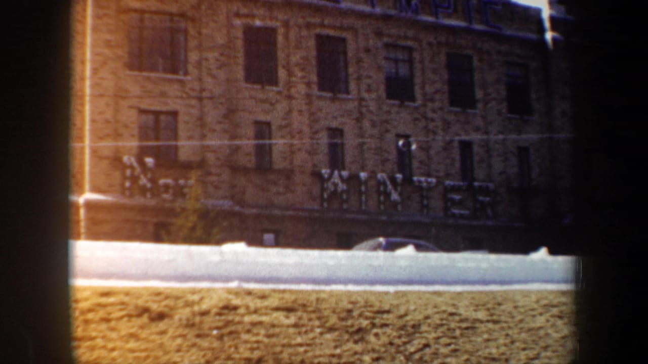 1962 New York home-movie frame of a brick facade, snowy curb, parked car, and large WINTER or OLYMPIC-style lettering.