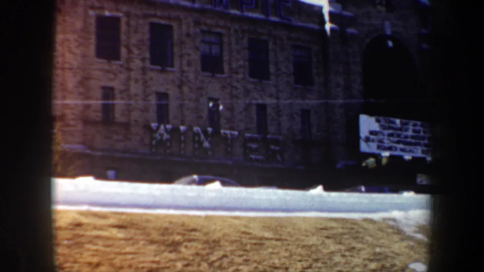 1962 New York 8mm frame showing a snowy street, brick building, large facade lettering, and a white sign near a dark entrance.